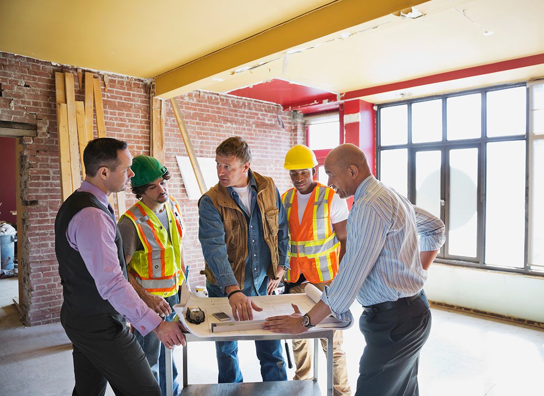 Contact - Team of Contractors Having a Meeting with Two Businessmen in a Building Under Construction Showing the Group the Building Blueprints