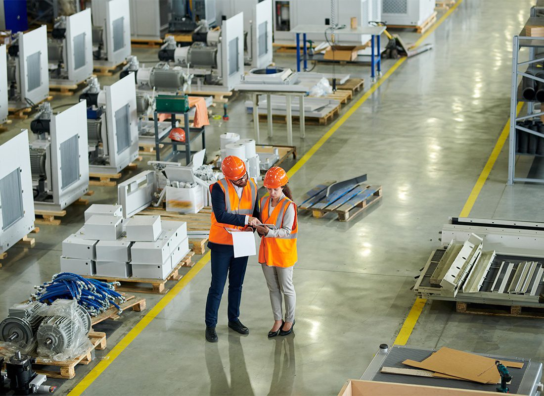 Insurance by Industry - Aerial View of Two Employees Standing in a Manufacturing Facility Full of Parts and Components Reviewing Documents