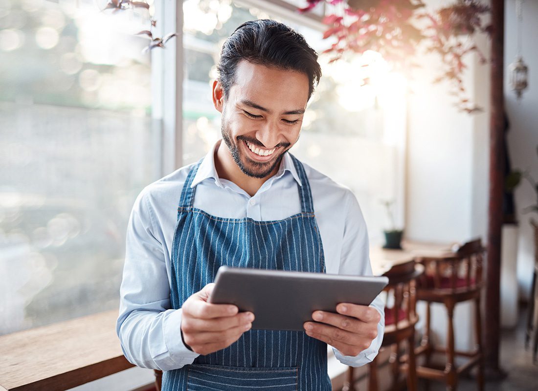 Blog - Closeup Portrait of a Cheerful Young Male Small Business Owner Wearing an Apron Reading on a Tablet While Standing Inside his Restaurant